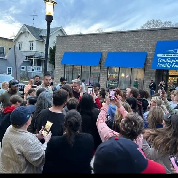 A crowd of people, many holding up phones, gathered around Gordon Ramsay outside The Neighborhood Kitchen.