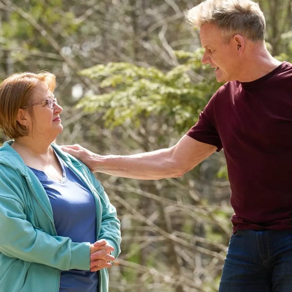 A man in a maroon shirt places his hand on a woman's shoulder, both smiling outdoors.