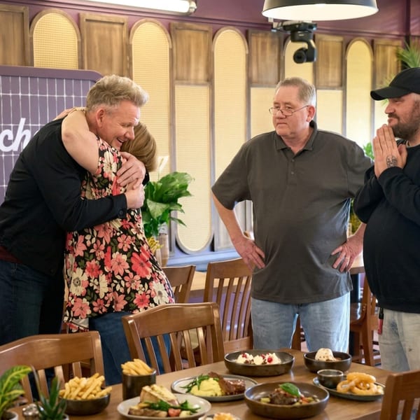 Gordon Ramsay hugs a woman in a floral dress, with two men and American dishes on a table.