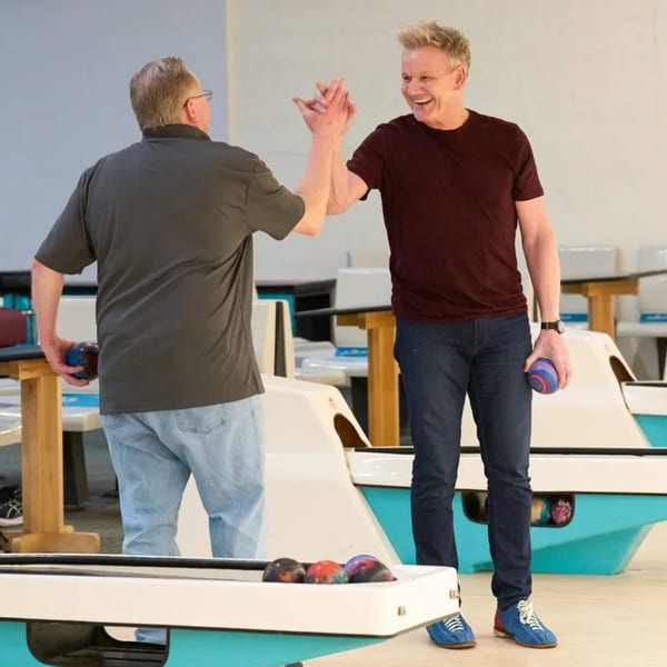 Two men high-fiving at a bowling alley, holding bowling balls, with a happy expression.