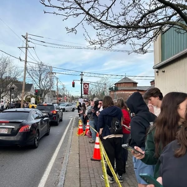 A long line of people waiting outside The Neighborhood Kitchen, marked by orange cones and yellow tape.