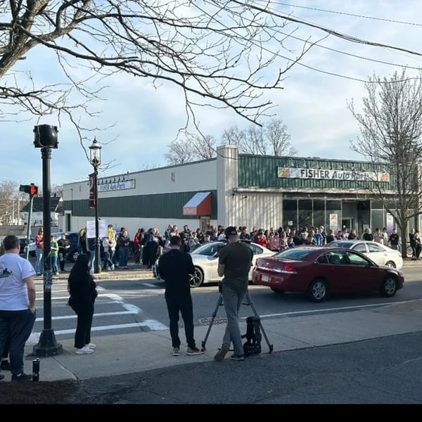 A crowd of people gathered outside Fisher Auto Parts, with cars and a camera crew in the foreground.