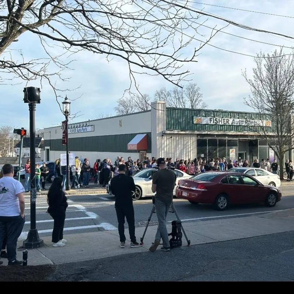 A crowd of people gathered outside Fisher Auto Parts, with cars and a camera crew in the foreground.