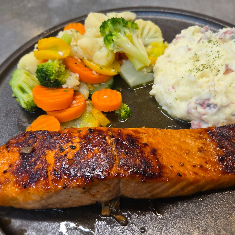 Glazed grilled salmon served with steamed broccoli, carrots, cauliflower, and a creamy herb potato salad on a dark plate.